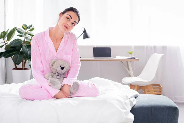 Attractive woman in pink pajamas with teddy bear sitting on bed at home — Stock Photo