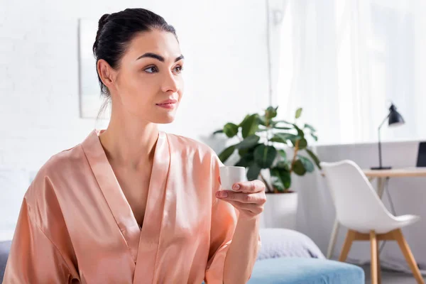 Retrato de mujer pensativa en albornoz de seda con taza de té caliente en la mano en la mañana en casa - foto de stock