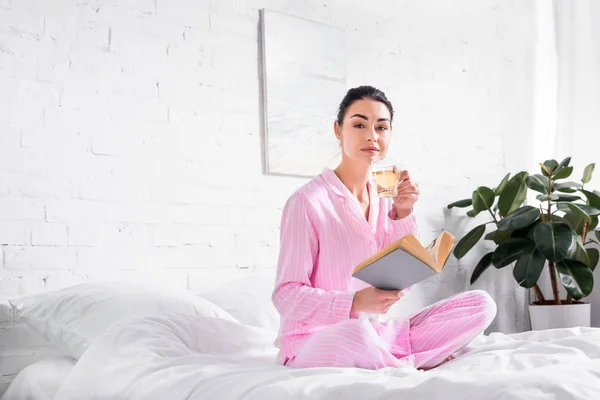 Женщина в пижаме с чашкой горячего чая и книги сидя на кровати утром дома — стоковое фото