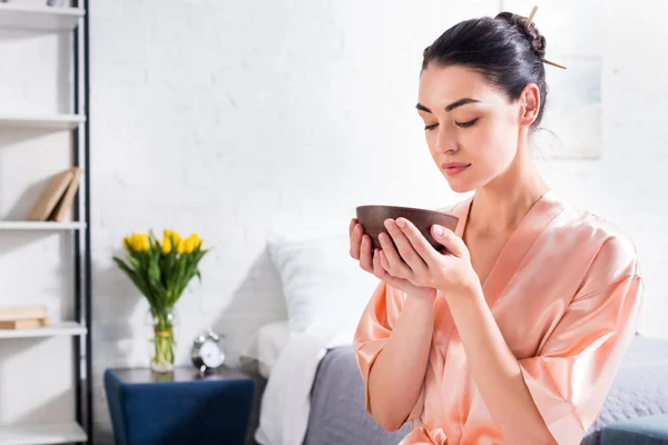 Mujer en albornoz de seda con cuenco de madera en las manos teniendo ceremonia del té en la mañana en casa - foto de stock