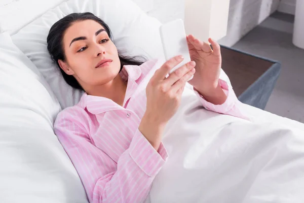 Portrait of woman in pajamas using smartphone while resting in bed at home — Stock Photo