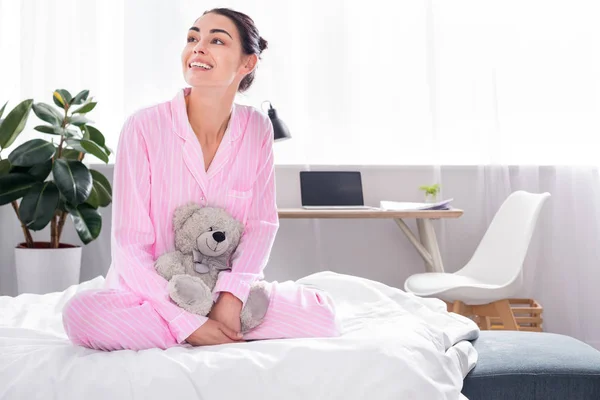 Cheerful woman in pink pajamas with teddy bear sitting on bed at home — Stock Photo