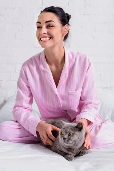 Веселая женщина в пижаме с британским короткометражным котом, отдыхающим на кровати дома — стоковое фото