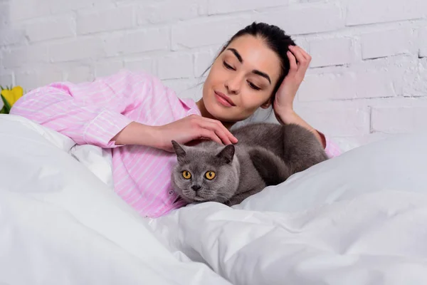 Ritratto di donna sorridente in pigiama che accarezza il gatto pantaloncini britannico mentre riposa sul letto a casa — Foto stock