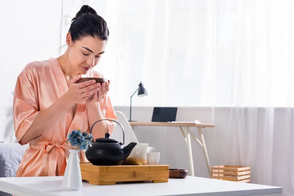 Mujer en albornoz de seda con cuenco de madera en las manos teniendo ceremonia del té en la mañana en casa - foto de stock