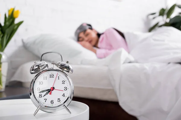 Foco seletivo de relógio e mulher em pijama rosa e máscara de dormir dormindo na cama em casa — Fotografia de Stock