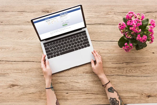 Vista parziale della donna al tavolo con computer portatile con sito facebook e impianto kalanhoe in vaso da fiori — Foto stock