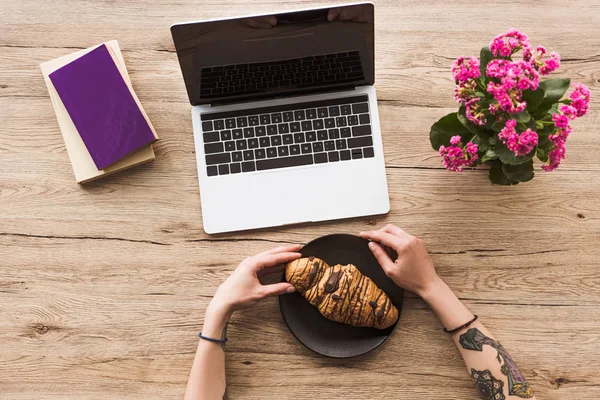 Colpo ritagliato di donna sul posto di lavoro con computer portatile, libri e croissant sul piatto — Foto stock