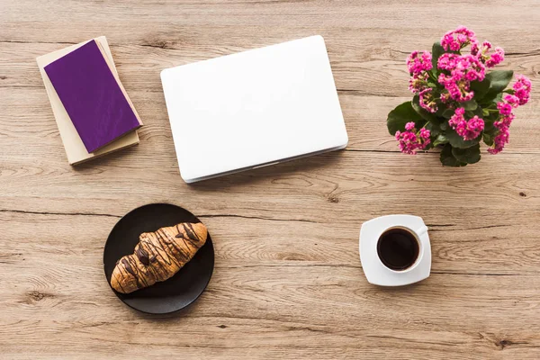 Posa piatta con computer portatile, libri, tazza di caffè, croissant sul piatto e pianta kalanchoe su tavolo in legno — Foto stock