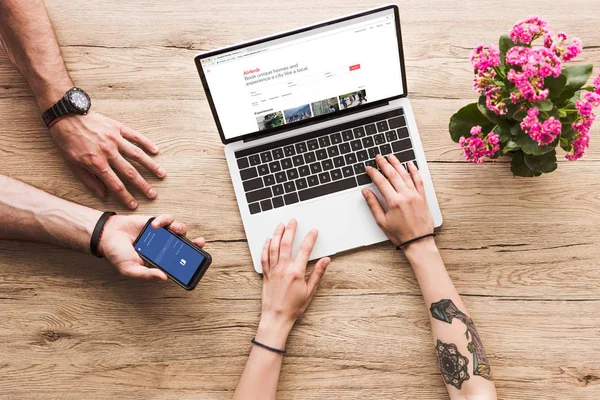 Tiro recortado de homem com smartphone com logotipo do facebook na mão e mulher na mesa com laptop com site airbnb e flor kalanchoe — Fotografia de Stock