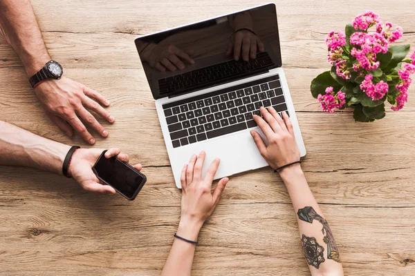 Cropped shot of man with smartphone in hand and woman at tabletop with laptop and kalanchoe flower — Stock Photo