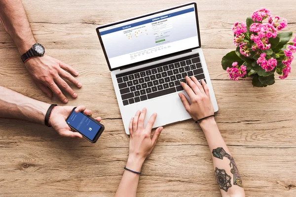 Cropped shot of man with smartphone with facebook logo in hand and woman at tabletop with laptop facebook website and kalanchoe flower — Stock Photo