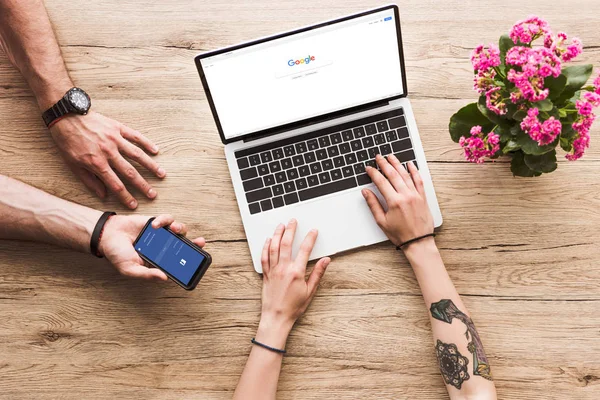 Cropped shot of man with smartphone with facebook logo in hand and woman at tabletop with laptop with google logo and kalanchoe flower — Stock Photo