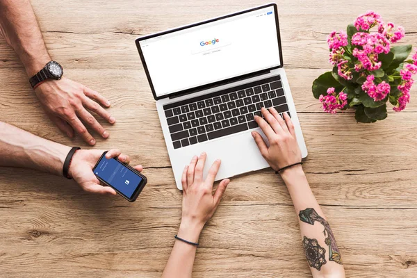 Recortado disparo de hombre con teléfono inteligente con el logotipo de facebook en la mano y la mujer en la mesa con el ordenador portátil con el logotipo de Google y la flor kalanchoe - foto de stock