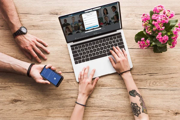 Abgeschnittene Aufnahme eines Mannes mit Smartphone mit Facebook-Logo in der Hand und einer Frau am Tisch mit Laptop mit linkedin Website und Kalanchoe-Blume — Stockfoto