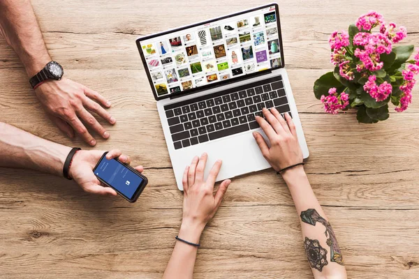 Schnappschuss eines Mannes mit Smartphone mit Facebook-Logo in der Hand und einer Frau am Tisch mit Laptop mit Pinterest-Website und Kalanchoe-Blume — Stockfoto