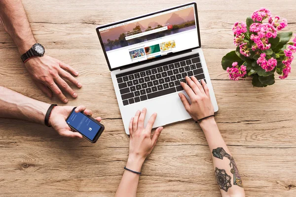 Recortado disparo de hombre con teléfono inteligente con el logotipo de facebook en la mano y la mujer en la mesa con el ordenador portátil con shutterstock sitio web y flor kalanchoe - foto de stock