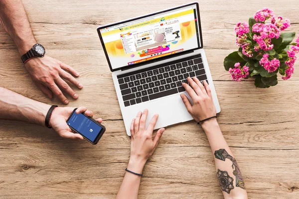 Cropped shot of man with smartphone with facebook logo in hand and woman at tabletop with laptop with aliexpress website and kalanchoe flower — Stock Photo