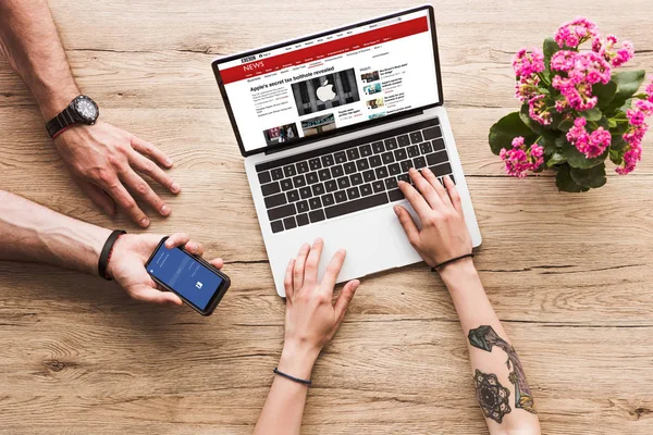 Corte tiro do homem com smartphone com o logotipo do facebook na mão e mulher na mesa com laptop com o site bbc e kalanchoe flor — Fotografia de Stock