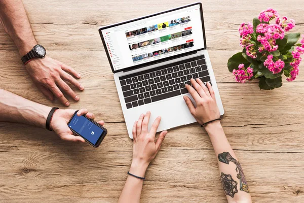 Cropped shot of man with smartphone with facebook logo in hand and woman at tabletop with laptop with youtube website and kalanchoe flower — Stock Photo