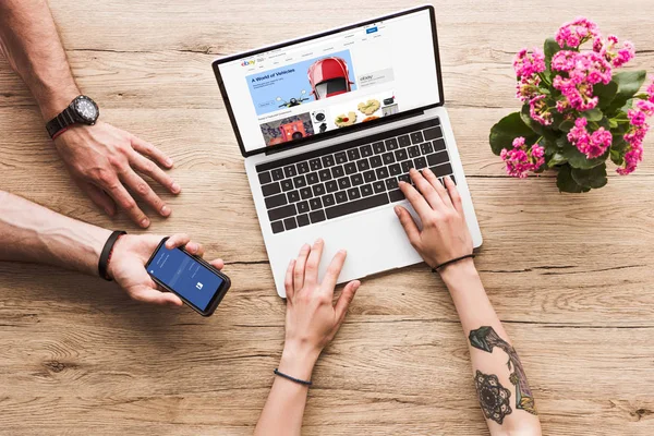 Cropped shot of man with smartphone with facebook logo in hand and woman at tabletop with laptop with ebay website and kalanchoe flower — Stock Photo