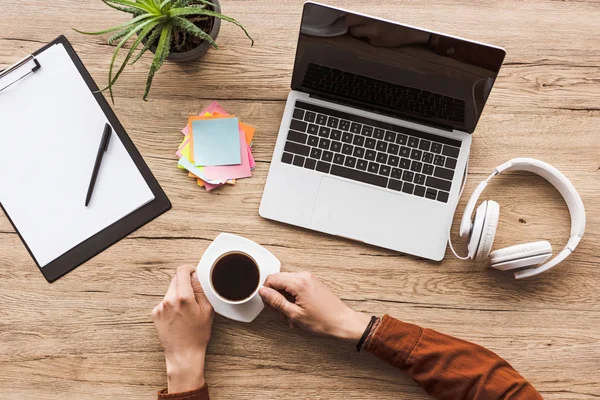 Tiro recortado de homem com xícara de café no local de trabalho com laptop, fones de ouvido, bloco de notas em branco e notas — Fotografia de Stock