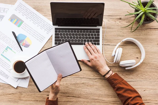 Cropped shot of man with empty notebook at workplace with headphones, papers and laptop — Stock Photo