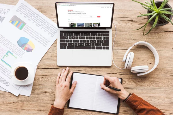 Teilansicht eines Mannes, der am Arbeitsplatz Notizen in Notizbuch mit Laptop, Airbnb-Website, Papieren, Tasse Kaffee und Kopfhörern macht — Stockfoto