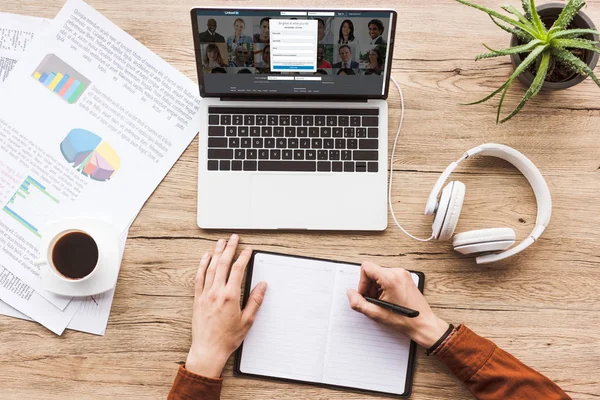 Partial view of man making notes in notebook at workplace with laptop with linkedin logo, papers, cup of coffee and headphones — Stock Photo