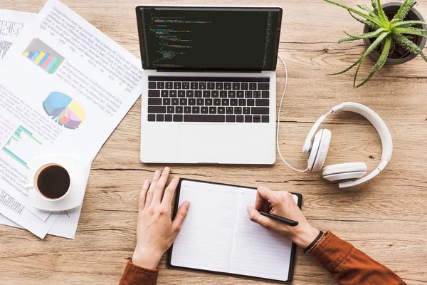 Teilbild eines Mannes, der am Arbeitsplatz mit Laptop, Papier, Tasse Kaffee und Kopfhörer Notizen in Notizbuch macht — Stockfoto