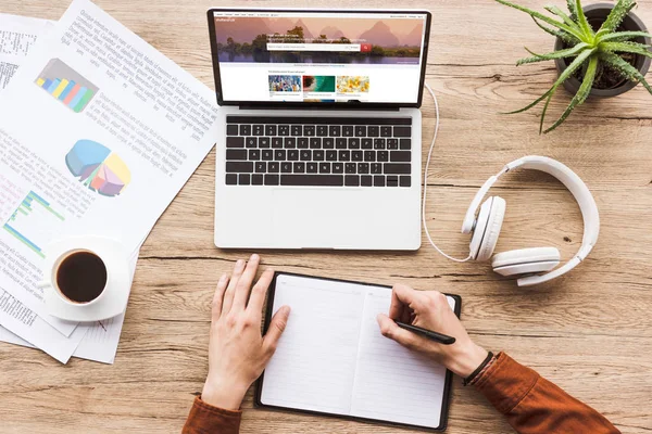 Частичный вид человека, делающего заметки в ноутбуке на рабочем месте с ноутбуком с Shutterstock сайт, бумаги, чашка кофе и наушники — стоковое фото