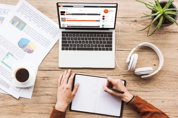 Partial view of man making notes in notebook at workplace with laptop with soundcloud website, papers, cup of coffee and headphones — Stock Photo