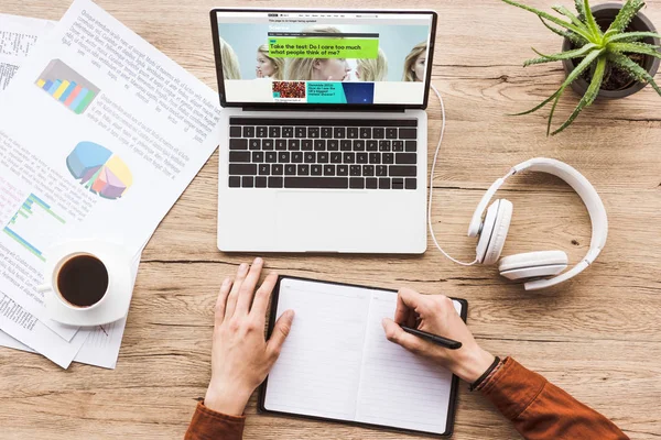 Partial view of man making notes in notebook at workplace with laptop with bbc website, papers, cup of coffee and headphones — Stock Photo