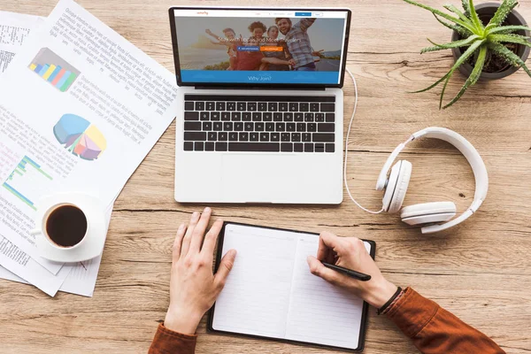 Teilansicht eines Mannes, der am Arbeitsplatz Notizen in Notizbuch mit Laptop mit Couchsurfing-Logo, Papieren, Kaffeetasse und Kopfhörern macht — Stockfoto