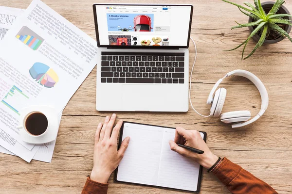 Imagen recortada del hombre que trabaja en la mesa con el ordenador portátil con el logotipo de ebay, auriculares, libro de texto, pluma, infografías, taza de café y planta en maceta - foto de stock