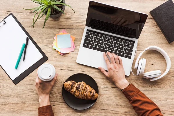 Cropped image of man working at table with laptop, headphones, textbook, clipboard, post it, potted plant, coffee cup and croissant — Stock Photo