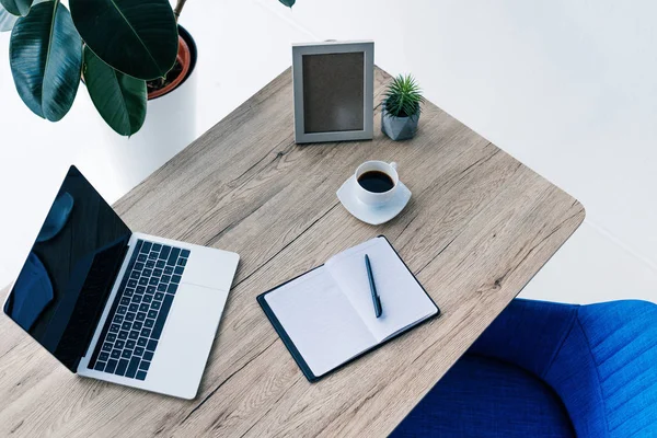 Elevated view of laptop with blank screen, empty textbook, photo frame, potted plant and coffee cup on table — Stock Photo