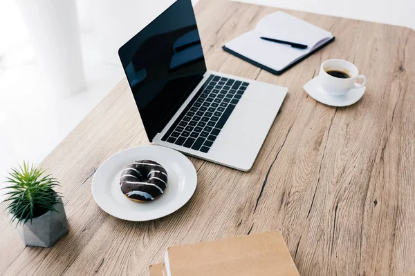 Closeup view of laptop with blank screen, doughnut, coffee cup, potted plant and textbooks — Stock Photo