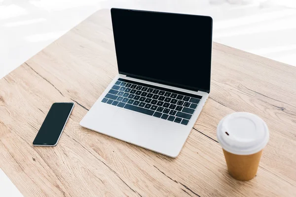 Vista de primer plano del teléfono inteligente con pantalla en blanco, ordenador portátil y taza de café en la mesa de madera - foto de stock