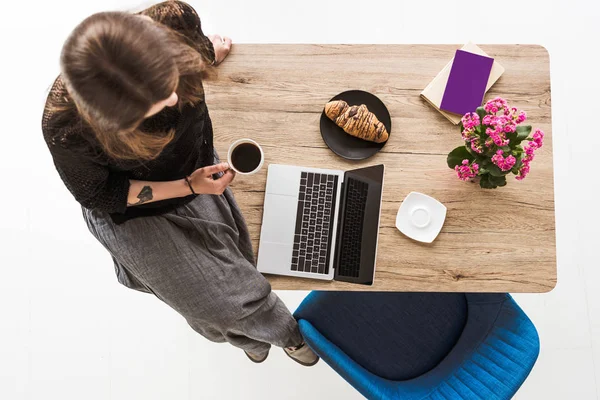 Vista dall'alto della donna con tazza di caffè seduta sul tavolo con croissant, computer portatile, fiori, libro e libro di testo — Foto stock