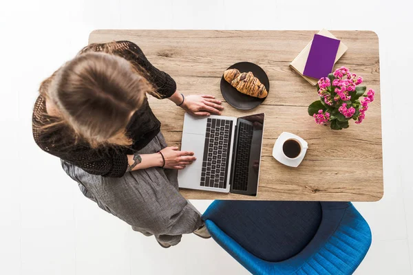 Vista aérea de la mujer escribiendo en el ordenador portátil con pantalla en blanco en la mesa con croissant, café, flores, libro y libro de texto - foto de stock