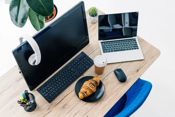 Vista ad alto angolo delle cuffie sul monitor del computer vuoto, laptop con schermo vuoto, cancelleria, croissant e tazza di caffè sul tavolo — Foto stock