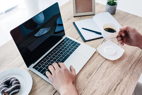 Image recadrée de l'homme avec tasse à café et beignet à table avec ordinateur portable, manuel, plante en pot et cadre photo — Photo de stock