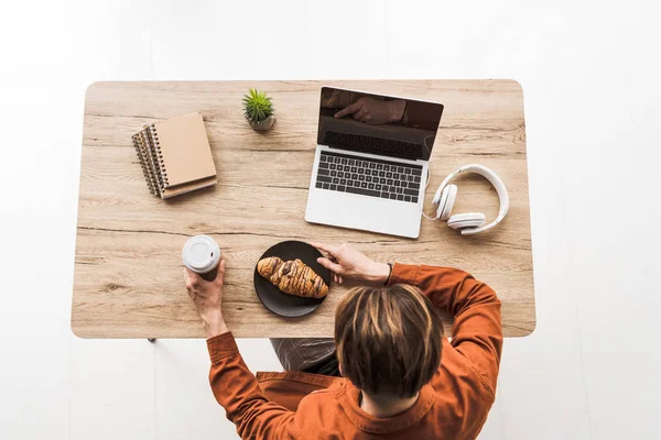 Повышенный вид человека с кофе и круассаном, работающего за столом с ноутбуком, наушниками, учебниками и горшком — стоковое фото