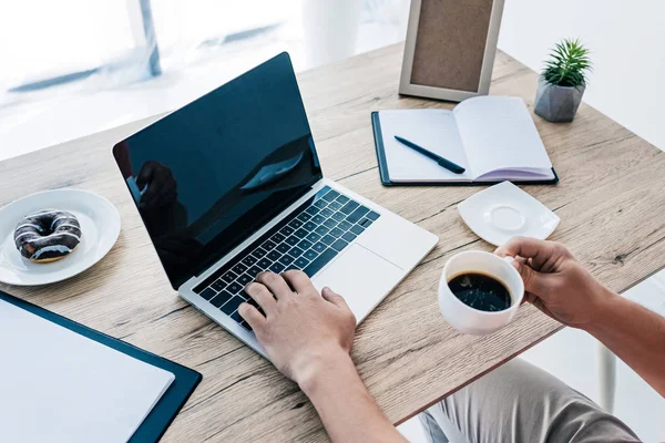 Imagen recortada del hombre con taza de café y rosquilla trabajando en la mesa con el ordenador portátil, portapapeles, libro de texto y marco de fotos — Stock Photo