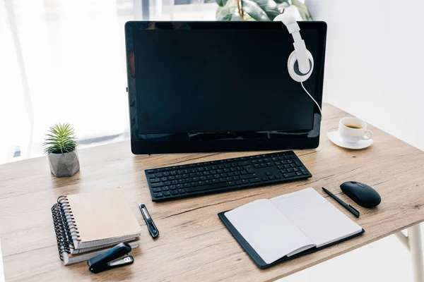 Vista de primer plano de los auriculares en el monitor de la computadora, libro de texto vacío, cuchillo de papelería, grapadora, taza de café y planta en maceta - foto de stock