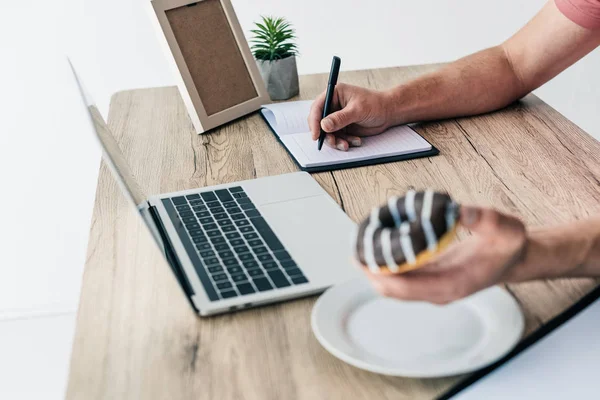 Cropped shot of man with doughnut writing in textbook at table with laptop, photo frame and potted plant — Stock Photo