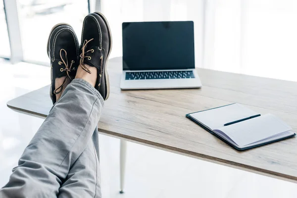 Частковий вигляд чоловічих ніг на столі з підручником і ноутбуком з порожнім екраном — стокове фото