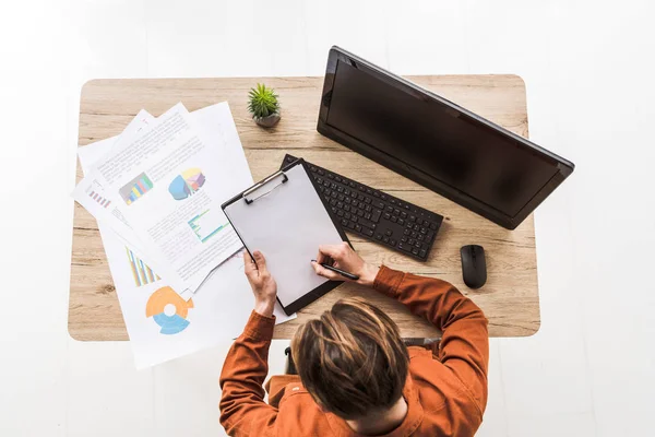 Vista elevada do homem que escreve na área de transferência vazia na tabela com infográficos, planta potted, computador, rato do computador e teclado — Fotografia de Stock