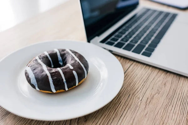 Closeup view of doughnut on plate and laptop with blank screen on wooden table — Stock Photo